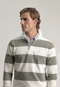 Camisa polo de manga larga con franjas alternas en verde oliva y blanco, con cuello de placket y un sutil logo bordado. Material de algodón.
