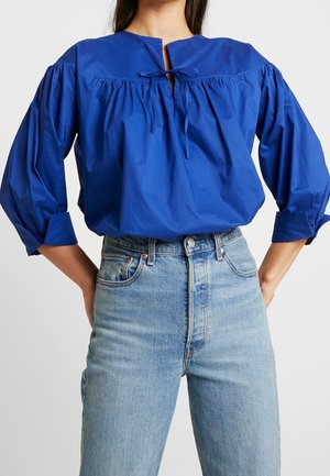 Kobieta ubrana w królewsko niebieską bluzkę z długim rękawem, ozdobioną marszczeniem i wiązanym detalem, w połączeniu z wysokim stanem wytartych niebieskich dżinsów.