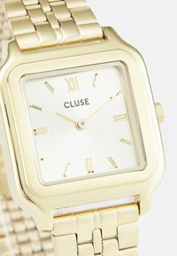 Montre carrée dorée avec un cadran blanc, des chiffres romains, et un bracelet en mailles. Présente un design minimaliste avec une finition brillante.