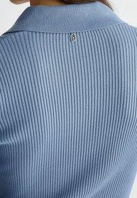 Ribbad ljusblå stickad topp med klassisk krage och en liten logodetalj vid halsen, som framhäver dess texturerade tyg och design.