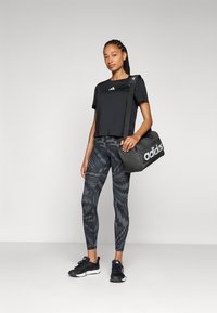 Sort cropped t-shirt med hvidt logo, mønstrede sorte leggings og sorte træningssko. Holder en mørkegrå taske med logo.
