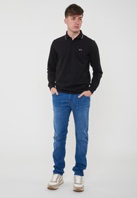 Polo a maniche lunghe nera con colletto bianco e logo verde, abbinata a jeans azzurri chiari e sneakers beige/bianco.