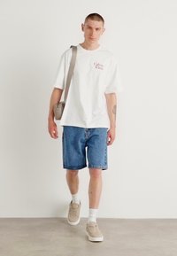 Witte katoenen t-shirt met rood 'Calvin Klein' logo, blauwe denim shorts, beige sneakers en een kleine grijze schoudertas.