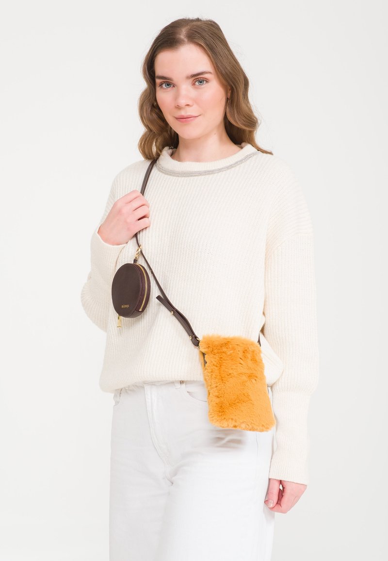 Brown leather circular bag with gold zipper and strap, paired with a fluffy bright orange crossbody pouch. Model wears a cream sweater.