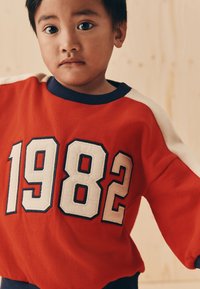 Sweat-shirt rouge avec grand imprimé blanc "1982", col bleu marine et accents crème aux épaules, doté d'une texture douce et d'une coupe décontractée.