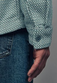 Light blue textured jacket with a woven pattern, featuring a buttoned cuff and visible stitching. Denim fabric is shown beneath.