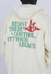 Sudadera crema con un gran gráfico en la parte posterior. El texto dice: "RESISTE SU CONTROL, ES TU LEGADO." Presenta una ilustración de una mano roja.