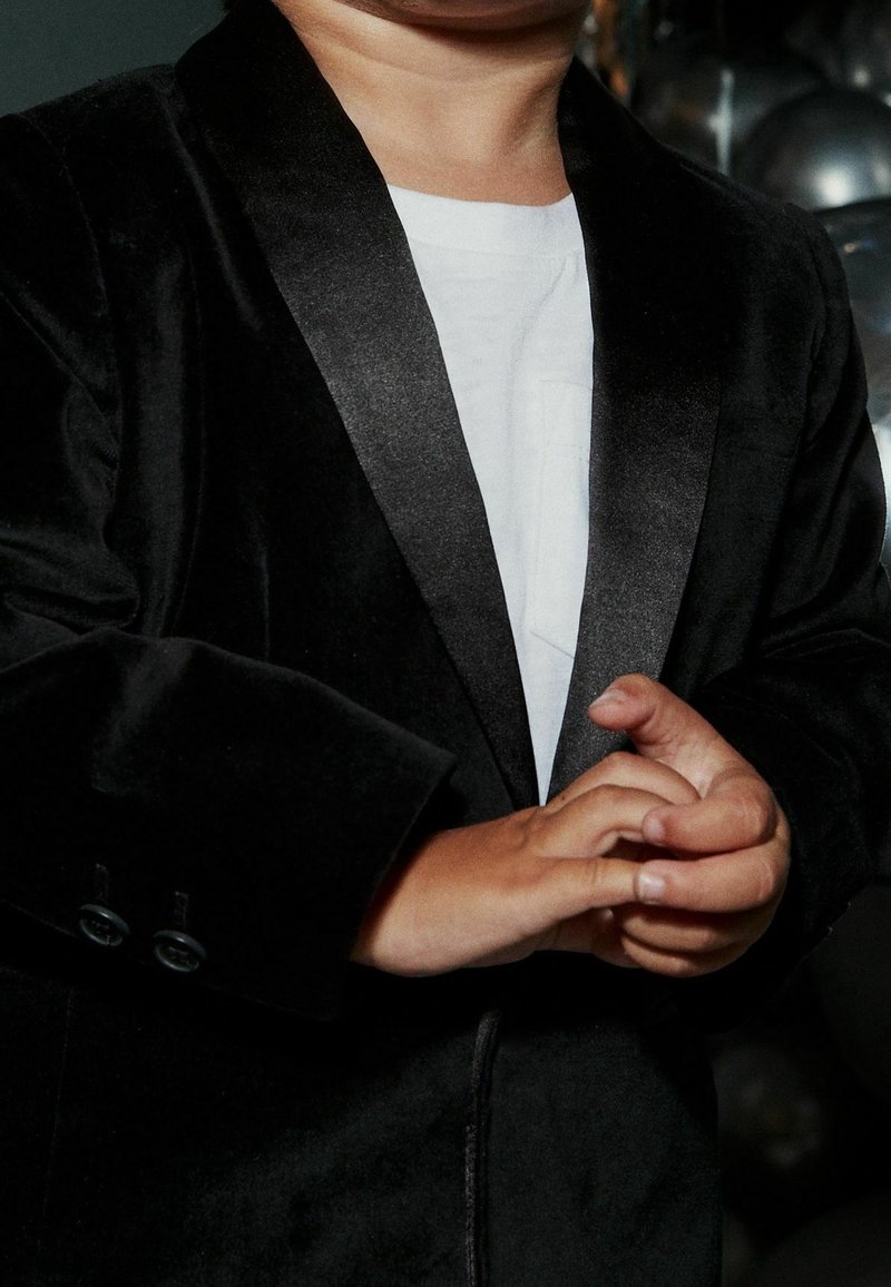 Blazer di velluto nero con rever in satin, indossato sopra una camicia bianca. Mani visibili unite, con la pelle liscia e le unghie corte.