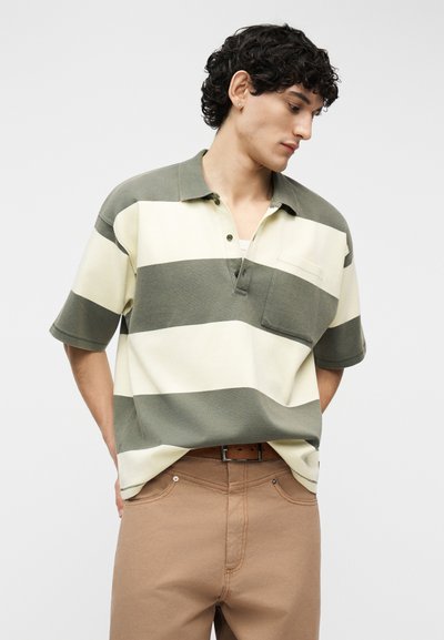 Νέος άνδρας που κοιτάζει προς τα κάτω, φορώντας μια κοντομάνικη, φαρδιά ράγκμπι πουκάμισο με πλατιές πράσινες και κρεμ ρίγες και μπεζ παντελόνι με καφέ ζώνη.