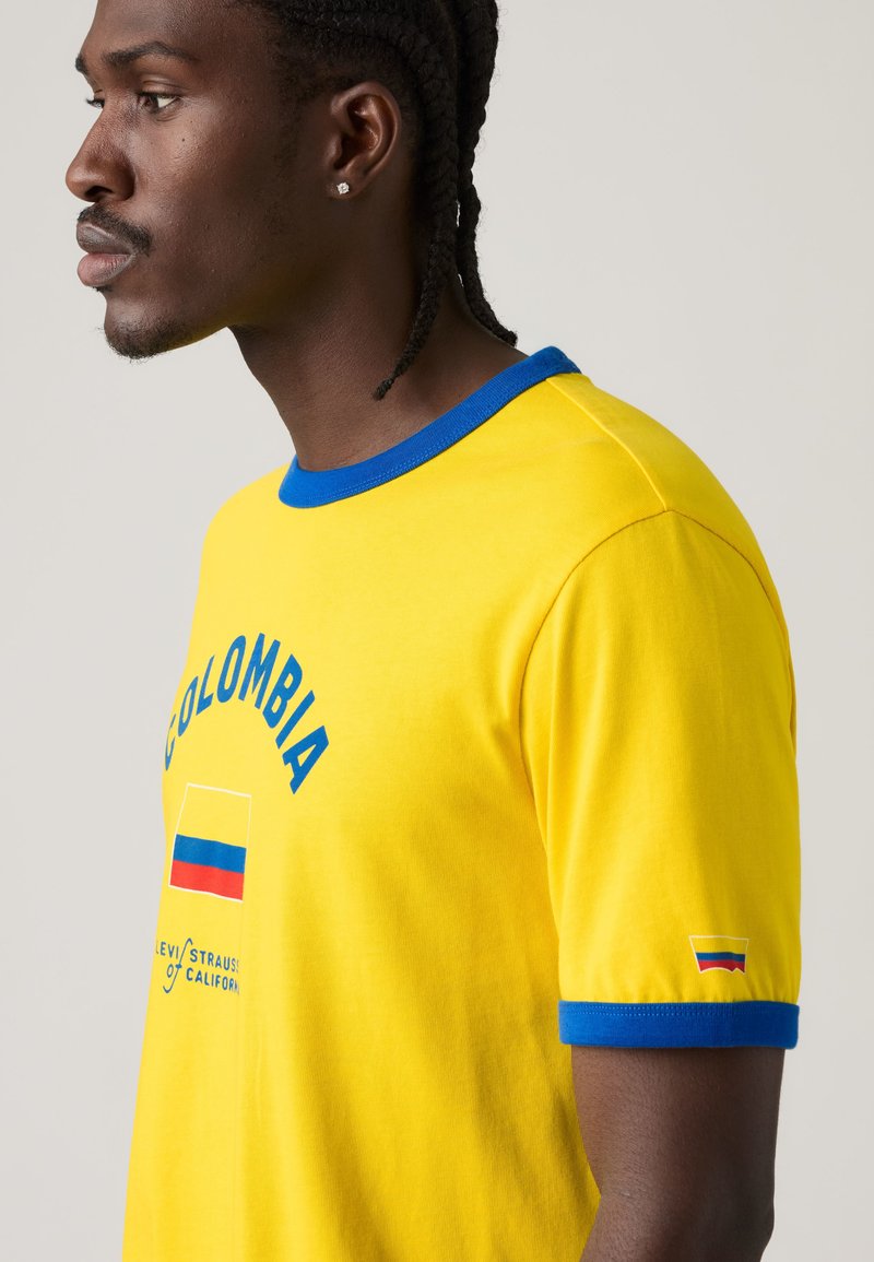 Homme aux cheveux tressés portant un T-shirt jaune avec col et bordures de manches bleus, avec le texte Colombie et des graphismes du drapeau.