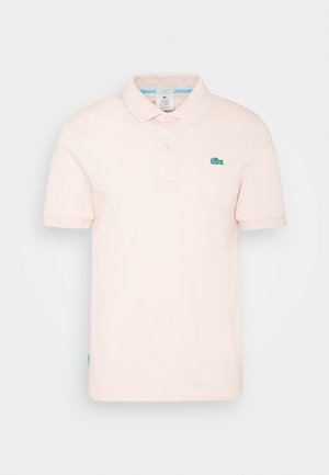 Mand iført en lyserød polo, off-white bukser og hvide sneakers med blå og gule detaljer, står foran en lysegrå baggrund.