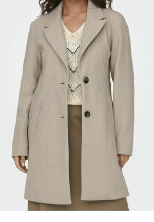 Mujer con abrigo de lana beige con cuello solapa, dos botones negros, bolsillos inclinados, sobre un suéter de punto crema y falda marrón.