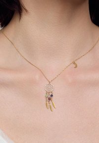 Złoty naszyjnik z okrągłym wisiorkiem w kształcie łapacza snów, ozdobionym wiszącymi kolorowymi koralikami i akcentami w postaci liści, leżący na skórze pod obojczykiem.