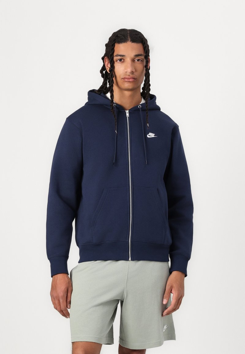 Marineblå zip-up hettegenser med snøring, hvit Nike-logo på brystet, frontlommer, ribbede mansjetter og nederkant. Kombinert med lysegrønne shorts.