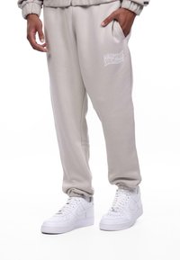 Light beige sweatpants with an elastic waist, side pockets, and white graphic text. Paired with white Nike Air Force 1 sneakers.