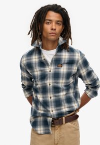 Camisa de franela a cuadros azul y crema con mangas largas, frontal abotonado y un bolsillo en el pecho que presenta un parche con logo. Lleva puesta una camiseta blanca debajo.