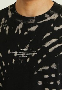 Černé a šedé tričko s efektom tie-dye s bílým logem Chasin' a textem umístěným uprostřed pod výstřihem na horní části hrudi člověka.