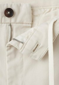 Zbliżenie na beżowe spodnie z ciemnym guzikiem, częściowo rozpiętym zamkiem błyskawicznym oraz białym sznurkiem przewleczonym przez dziurkę na guzik.