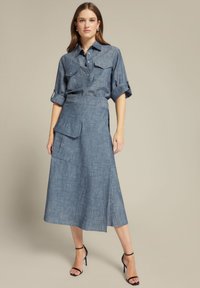 Denim shirt met opgerolde mouwen, dubbele borstzakken; gecombineerd met een lange, gestructureerde denim rok met een zijzak en split. Zwarte hakken met banden.
