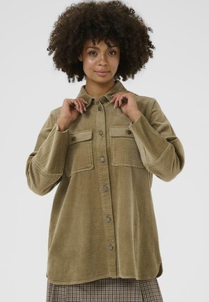 Frau mit lockigen Haaren, die eine geknöpfte khakifarbene Cordjacke mit zwei Brusttaschen trägt und den Kragen mit beiden Händen hält.