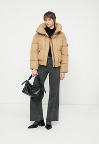 Sieviete, tērpusies brūnā pūkainā jaku, melnā turtleneck blūzē, pelēkās platās biksēs, melnās smailes kurpes, tur melnu rokas somu uz baltā fona.
