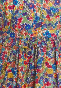 Bunt gemustertes Blumenkleid mit einem Rüschensaum, das eine Mischung aus roten, blauen, gelben und grünen Blumen auf einem weißen Hintergrund zeigt. Sichtbares Knopfdetail.