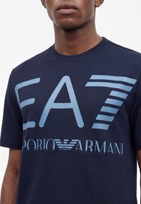 Νέος άνδρας φοράει μπλε σκούρο T-shirt Emporio Armani EA7 με μεγάλο ανοιχτό μπλε λογότυπο και κείμενο στο στήθος.