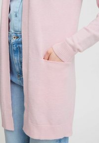 Cardigan a maglia rosa chiaro con maniche lunghe, polsini a coste, apertura frontale e una tasca laterale. Indossato sopra una camicia blu e jeans di denim.