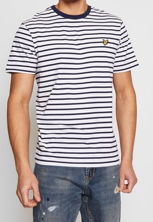 Man draagt een wit en marineblauw gestreept T-shirt met korte mouwen met een geel vogel-logo, gecombineerd met blauwe versleten spijkerbroek.