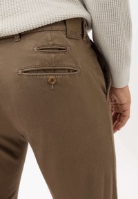 Bruine gestructureerde broeken met dubbele achterzakken, voorzien van een knoopsluiting en een klein merketiket, gecombineerd met een lichtgrijze geribbelde trui.