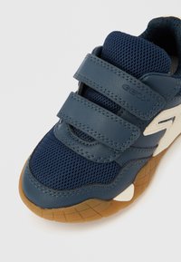 Σκούρο μπλε αθλητικό παπούτσι από διχτυωτό ύφασμα και δέρμα, με δύο λουριά Velcro, λαστιχένια εξωτερική σόλα και αντίθετες λευκές λεπτομέρειες.