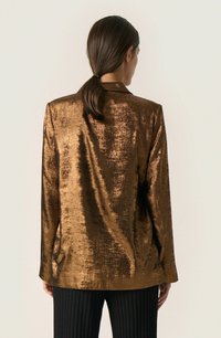 Bronzový metalický blazer s lesklým, strukturovaným povrchem, s jedním knoflíkem a klopovými límci, spárovaný s černými ribkovanými kalhotami.