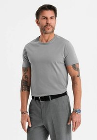 Maglietta di cotone grigia con scollo rotondo e maniche corte, indossata da un uomo. Presenta tatuaggi visibili su entrambe le braccia e una cintura nera.