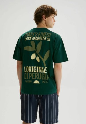 Jeune homme aux cheveux bouclés portant un t-shirt vert foncé avec un motif de branche d'olivier et un texte sur l'huile d'olive extra vierge italienne.