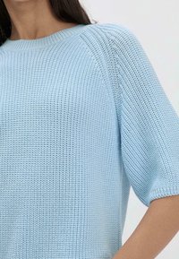 Femme portant un pull en maille bleu clair à manches courtes avec un col rond, montré des épaules jusqu'au milieu du bras sur un fond uni.