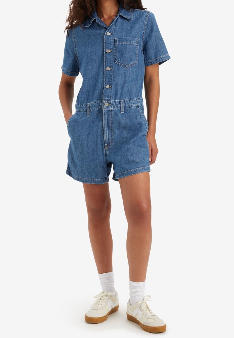 Femme portant une combinaison-short en denim bleu à manches courtes avec des poches avant, accompagnée de chaussettes blanches et de baskets crème.