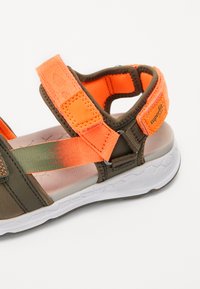 Rudos ir oranžinės spalvos sandalai su reguliuojamais Velcro diržais, tekstūruota medžiaga, paminkštintu padu ir baltu guminžemiu.
