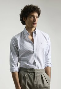 Camisa blanca de botones con rayas azul claro, corte entallado, mangas remangadas, combinada con pantalones gris de talle alto y plisados.
