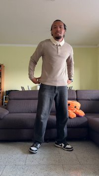 Uomo che indossa un polo beige a maniche lunghe e jeans neri, in piedi in un soggiorno con un divano grigio e un peluche di polpo arancione.
