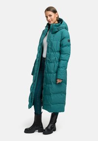 Tirkīzzila pūkļu mētelis ar kapuci, sānu kabatām un pogām priekšpusē; matēta apdare; savienots ar melniem potītes zābakiem un džinsiem.