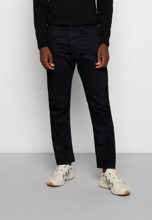 Σκούρο μπλε τζιν παντελόνι με στενή γραμμή, πέντε τσέπες και έναν διακριτικά υφασμένο φινίρισμα, συνδυασμένο με ανοιχτά μπεζ αθλητικά παπούτσια.