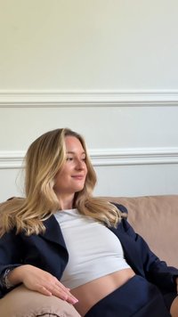 Szőke nő egy bézs kanapén ül, fehér crop topot és sötétkék blézert visel. Hosszú haja van és békés kifejezés az arcán.