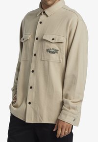 Button-up beige flannel shirt with two chest pockets, dark brown buttons, and embroidered "Billabong" logo. Soft textured fabric.