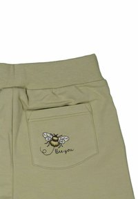 Beige puuvillapantsit, joissa on takatasku, jossa on brodeerattu mehiläissuunnittelu ja teksti "Ole oma itsesi" tyylikkäällä kursiivilla.