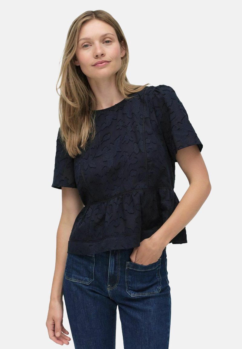 Tummansininen peplum-toppi, jossa on tekstuuri, lyhyet hihat ja pyöreä pääntie, yhdistetty tummiin farkkuihin.