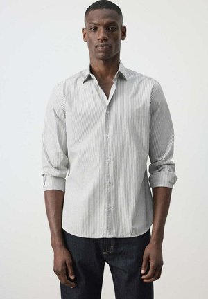 Hombre con camisa de botones de rayas finas en gris claro y blanco con las mangas remangadas y pantalones oscuros, de pie frente a un fondo blanco liso.