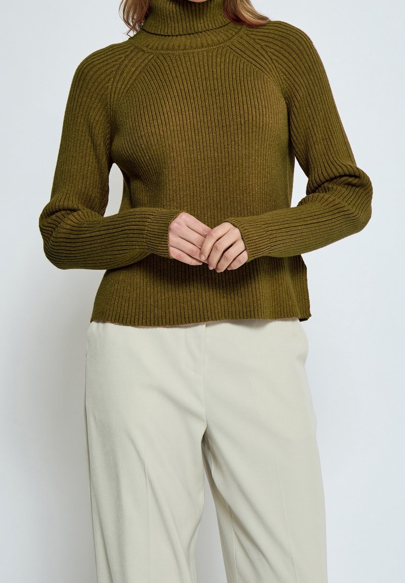 Femme portant un pull à col roulé côtelé vert olive et un pantalon écru, les mains délicatement jointes au niveau de la taille.