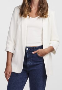 Blazer blanc ajusté avec des manches retroussées, associé à un haut blanc près du corps et un jean en denim bleu foncé taille haute.