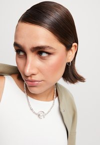 Collier en perles argentées avec un pendentif en perle, porté sur un haut blanc, associé à un cardigan vert clair. Cheveux lisses, maquillage neutre.