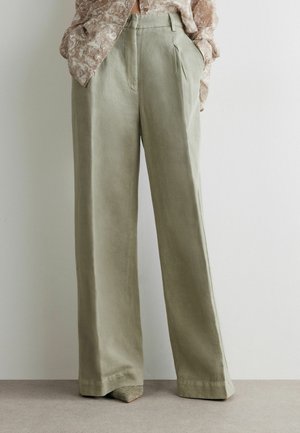 Frau trägt lockere, hoch taillierte, weit geschnittene Hosen in hellem Oliv mit seitlichen Taschen und eine gemusterte, langärmlige Bluse, die eingesteckt ist.